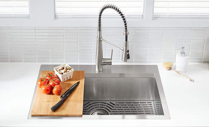 Bright natural light illuminates an overhead view of a stainless-steel Kohler kitchen sink with a cutting board resting over half of it. On the cutting board are five bright-red tomatoes and a small carton of white button mushrooms, along with black slicing knife. 