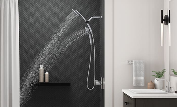 A Kohler dual-head shower fixture streams water in an empty, modern standing shower with dark gray tiling and a floating shelf with cleansing products resting on top.