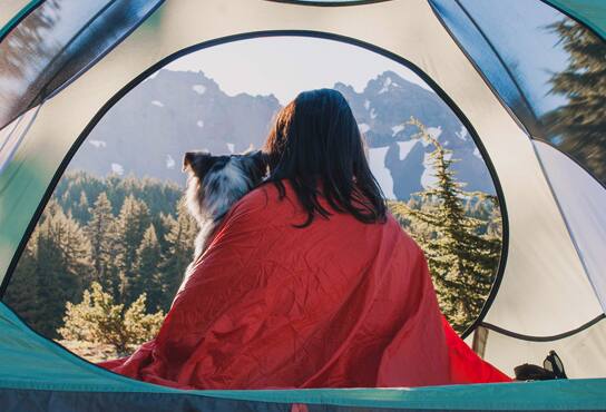 A woman and her dog sit in a tent, bundled up in a blanket, looking out at a forest.