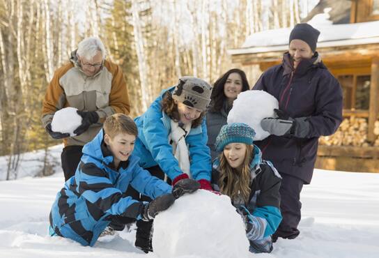 A large family sits outside their cabin, building a snowman and laughing.